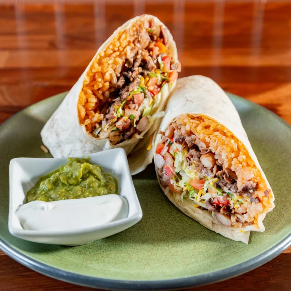 Two Miguelito's burritos, cut in half, revealing rice, meat, and vegetables, with guacamole and sour cream.