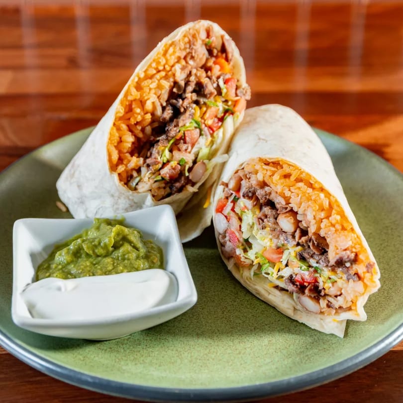 Two Miguelito's burritos, cut in half, revealing rice, meat, and vegetables, with guacamole and sour cream.