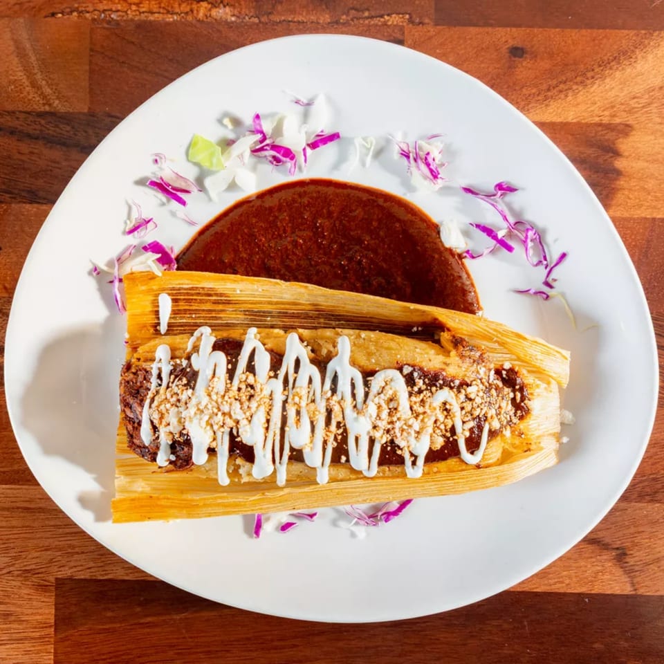 A Miguelito's Cocina Mexicana tamale with dark mole sauce, white drizzle, and crumbled nuts on a white plate.